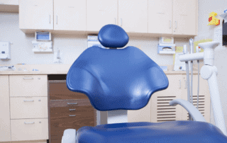 A blue dental chair is positioned in the center of a clean, modern dental practice, surrounded by light-colored cabinets, drawers, and various dental supplies.
