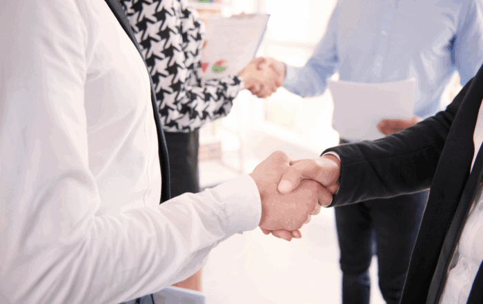 Close-up of two people in business attire shaking hands, with two others blurred in the background also shaking hands, suggesting a professional or business meeting.