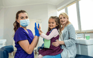 A female dental professional in scrubs and a mask gives a high-five to a smiling young girl sitting on her mother’s lap in a dental office. The mother and child both look happy and relaxed.