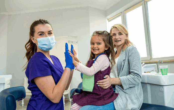 A female dental professional in scrubs and a mask gives a high-five to a smiling young girl sitting on her mother’s lap in a dental office. The mother and child both look happy and relaxed.